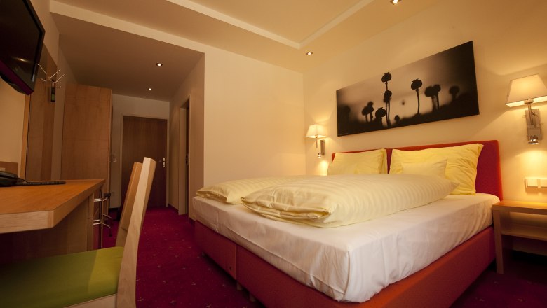 Faulenzerhotel Schweighofer, © Faulenzerhotel Schweighofer Ein gemütliches Hotelzimmer mit Doppelbett, Schreibtisch und Wandbild. Warme Beleuchtung und rote Teppiche schaffen eine einladende Atmosphäre.