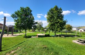 Spielplatz, © Berger Ein Spielplatz mit Klettergerüst, Schaukeln und Picknicktisch auf einer grünen Wiese unter blauem Himmel.