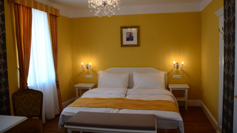 Kaiser Karl Zimmer, © Engelbrecht Elegantes Hotelzimmer mit gelben Wänden, Doppelbett, Kronleuchter und Bild an der Wand.