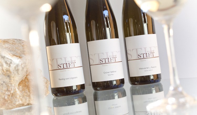 Wein ist Genuss, Leidenschaft und die Liebe zur Natur, © Astrid Bartl Drei Weinflaschen der Marke Stift auf einem Tisch, umgeben von Weingläsern.