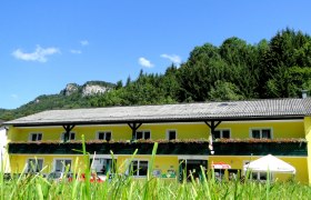 Voralpenhof, © Voralpenhof Gelbes Gebäude mit Blumenbalkon vor bewaldetem Hügel und blauem Himmel.