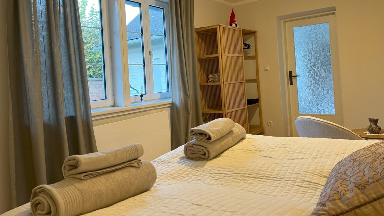 Schlafzimmer, © Waltraud Forstner Ein gemütliches Schlafzimmer mit einem Bett, auf dem Handtücher liegen, einem Fenster mit Vorhängen und einem Regal mit Dekoration.