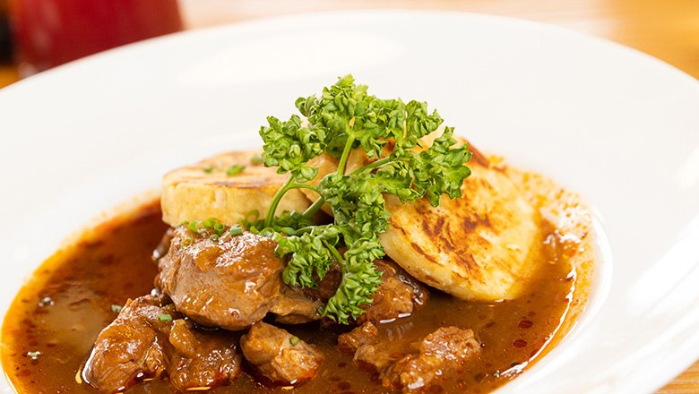 Eis-Greissler Kulinarik-Stadl Rindsgulasch, © Blochberger Eisproduktion GmbH Ein Teller mit Rindsgulsch und Knödeln mit Petersilie garniert.