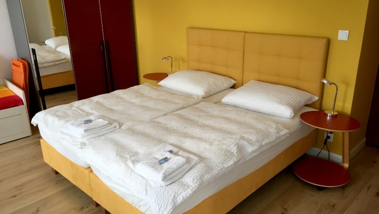 Doppelbett, © © E. Renfer Ein Doppelbett mit gelbem Kopfteil, weißen Bettdecken und Kissen in einem Zimmer mit gelber Wand und rotem Schrank.
