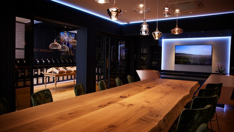 Weinlounge, © Ben Leitern Eleganter Raum mit langem Holztisch, modernen Lampen und einem Bildschirm an der Wand.