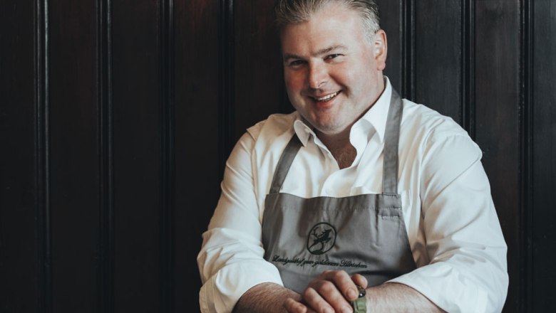 Stephan Solich verrät sein Wissen in Kochkursen, © Niederösterreich Werbung/David Schreiber Mann in weißem Hemd und Schürze lächelt vor dunkler Holzwand.