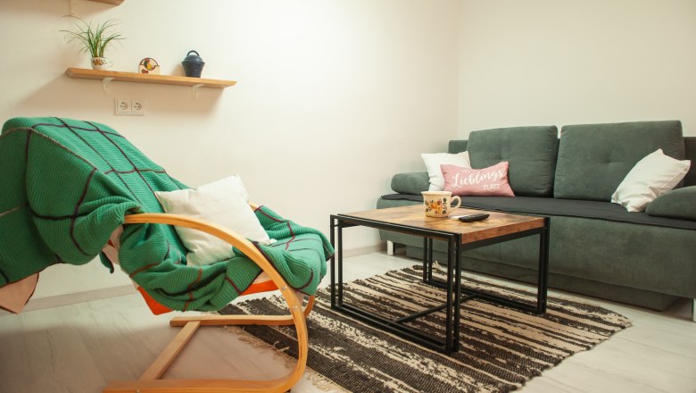 Wohnzimmer, © Fam. Heinrich Gemütliches Wohnzimmer mit grauem Sofa, Holztisch, grünem Schaukelstuhl und Regalen mit Pflanzen.