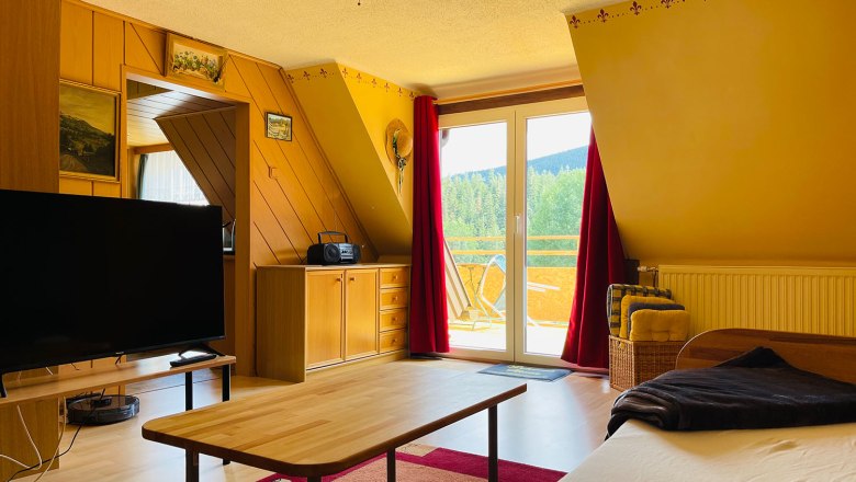 Wohnzimmer, © Angelika Burger Gemütliches Wohnzimmer mit Holzmöbeln, großem Fernseher, Tisch und Zugang zu einem Balkon mit Aussicht auf grüne Landschaft.