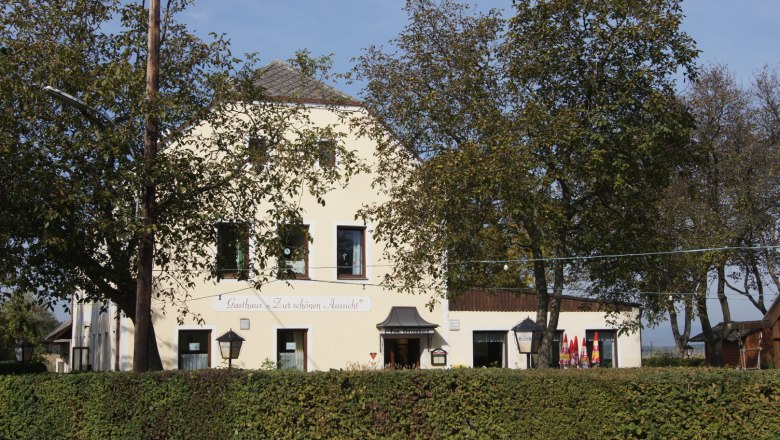 gasthaus-zur-schoenen-aussicht-1_2, © Familie Osterhaus Ein gelbes Gasthaus mit der Aufschrift 'Gasthaus Zur schönen Aussicht', umgeben von Bäumen und Hecken.