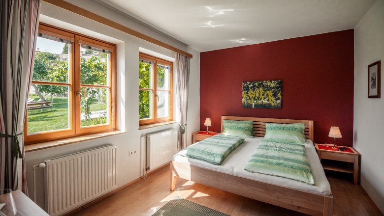 Gästehaus-Winzerhof Bernhard, © Jolly Schwarz Helles Schlafzimmer mit Doppelbett, roten Wänden und großen Fenstern mit Blick ins Grüne.
