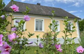 Herzlich Willkommen, © Waltraud Forstner Ein gelbes Haus mit weißen Fensterrahmen, umgeben von blühenden rosa Blumen im Vordergrund.