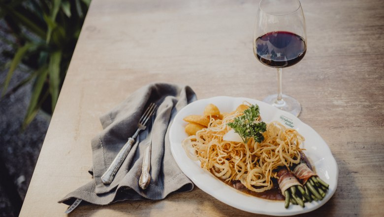 Zwiebelrostbraten, Kartoffeln, Speckbohnen, © Niederösterreich Werbung/Rita Newman Ein Teller mit Zwiebelrostbraten, Kartoffeln und Speckbohnen, daneben ein Glas Rotwein auf einem Holztisch.