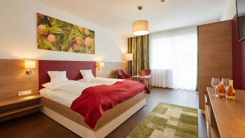Zimmerbeispiel im Weinhof Aufreiter, © Michael Schafranek Modern eingerichtetes Hotelzimmer mit Doppelbett, roter Decke, Holzmöbeln und großem Fenster. An der Wand hängt ein Bild mit Früchten.