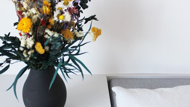 Gästezimmer, © Stadthotel Hollabrunn Ein Tisch mit einer schwarzen Vase voller bunter Blumen, einem blauen Magazin mit der Aufschrift 'HOLLA!' und einem weißen Kissen.