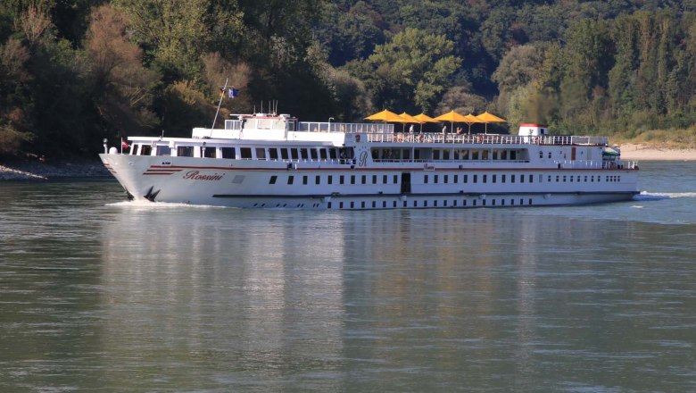 Donauschiff vor der Haustüre, © Szilágyi Ein Flusskreuzfahrtschiff namens Rossini fährt auf der Donau, umgeben von bewaldeten Ufern.