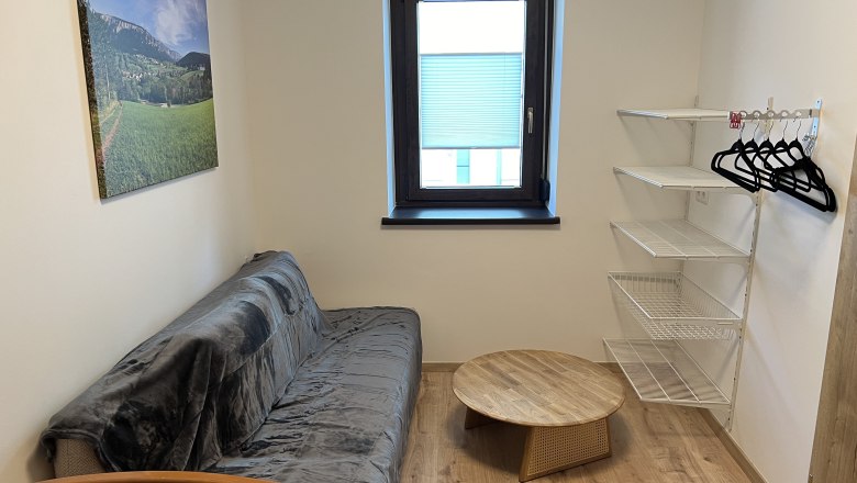 Zimmer, © Anton Sommer Kleines Zimmer mit Sofa, rundem Holztisch, Wandregal und Fenster.