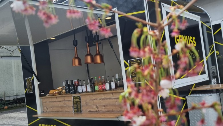 Unterwegs im Foody, © Die zwoa Weinviertla Ein schwarzer Foodtruck mit der Aufschrift 'Genuss Fahrbrik' steht im Freien. Im Vordergrund sind unscharfe Blüten zu sehen.