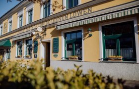 Aussenansicht Zum goldenen Löwen, © Familie Frey/Zum goldenen Löwen Außenansicht eines gelben Gebäudes mit der Aufschrift 'Zum goldenen Löwen'. Fenster mit Blumenkästen und grünen Markisen.