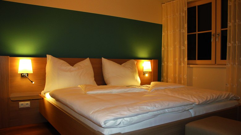 Gästezimmer, © Widhalm Ein gemütliches Gästezimmer mit einem Doppelbett, zwei Nachttischlampen und einem Fenster mit Vorhängen.