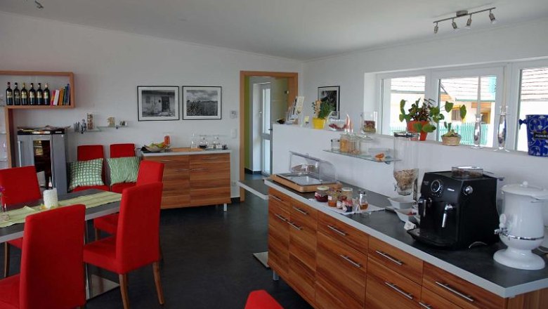 Holzheu Frühstücksraum, © Philipp Monihart Frühstücksraum mit roten Stühlen, Holzmöbeln und Kaffeemaschine.