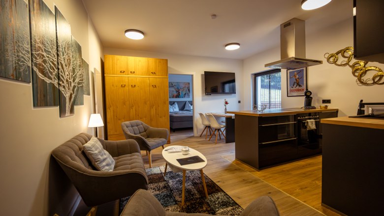 Ötscher Lodge, © Niederösterreich Werbung / Maximilian Pawlikowsky Modern eingerichtetes Wohnzimmer mit Küche, Sofa, Sesseln und Esstisch. Helle Holzmöbel und Wandkunst sind zu sehen.