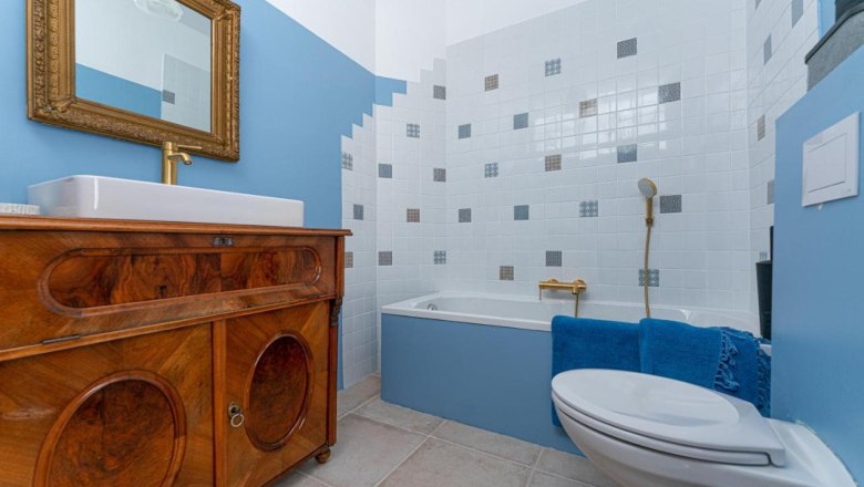 Badezimmer, © Guestia GmbH Ein Badezimmer mit blauen Wänden, einer Badewanne, einem Waschbecken auf einem Holzschrank und einem goldenen Spiegel.