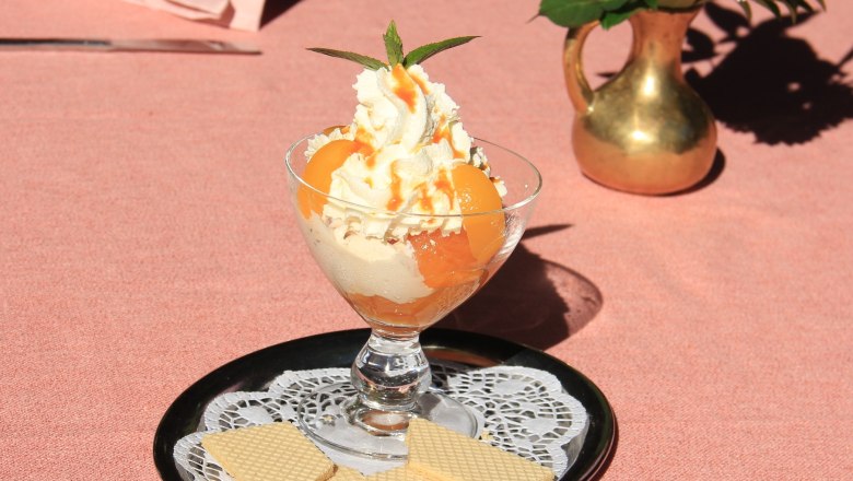 Marilleneistraum, © Schendl Ein Dessertglas mit Eiscreme, Sahne und Aprikosen auf einem Tablett mit Waffeln, daneben eine Vase mit Blumen.