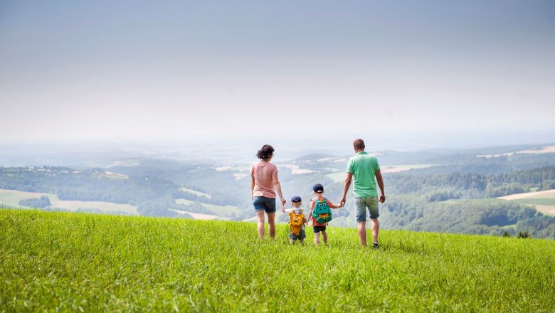 Bakabu Erlebnisweg in Hochneukirchen, © MG Hochneukirchen-Gschaidt, Stefan Knittel Familie spaziert über eine grüne Wiese mit weitem Blick auf die Landschaft.