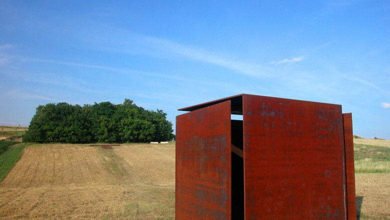 Windwürfelhaus, © StadtGemeinde Mistelbach Rostiger Metallwürfel auf einem Feld mit Bäumen im Hintergrund.