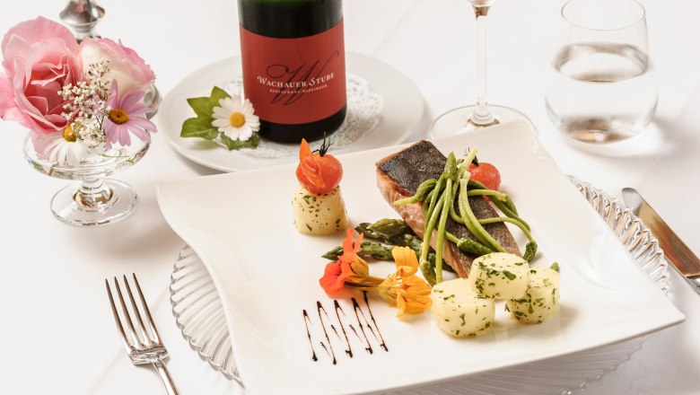 Wachauerstube, © Rita Newman Elegantes Gericht mit Fisch, Spargel und Kartoffeln auf weißem Teller, dekoriert mit Blumen. Im Hintergrund eine Flasche Wein und ein Glas.
