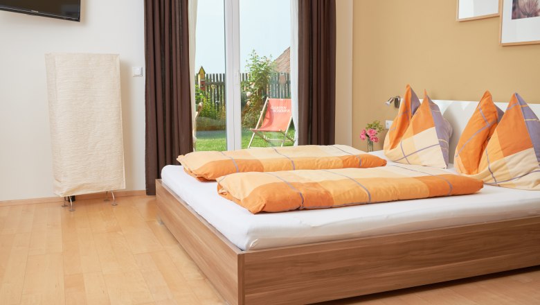 Holzheu Zimmer, © Philipp Monihart Ein gemütliches Schlafzimmer mit einem Doppelbett, orangen Bettdecken und einem Blick auf einen Garten durch eine Glastür.