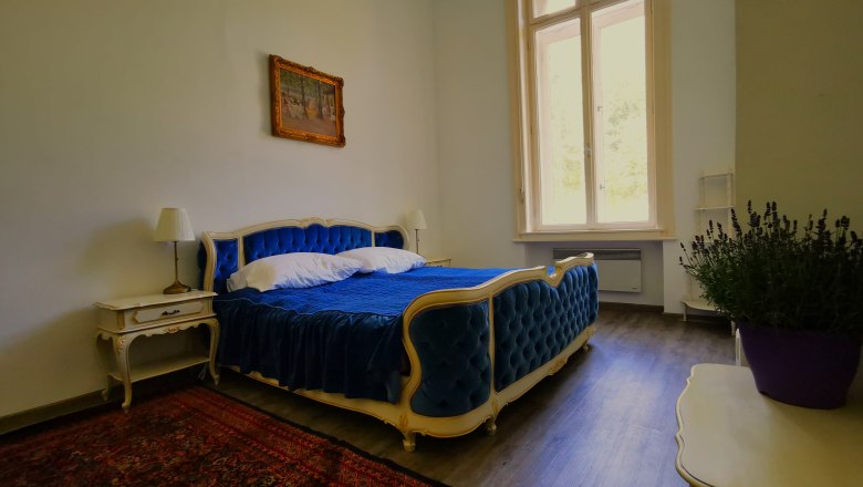 Schlafzimmer im Appartement Nikolaus, © Herrnhof Elegantes Schlafzimmer mit blauem Bett, antiken Möbeln und großem Fenster.