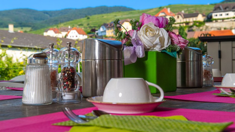 Frühstück im Hotel Weinquadrat, © ivan-lukacic-photography Frühstückstisch im Freien mit rosa Tischsets, Kaffeetassen, Salz- und Pfefferstreuern und Blumenarrangement, im Hintergrund eine malerische Landschaft.