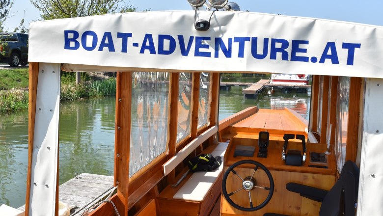 An Borad der Boat Adventure, © Boat Adventure Innenansicht eines Bootes mit Steuer und Sitz, beschriftet mit 'Boat-Adventure.at'.