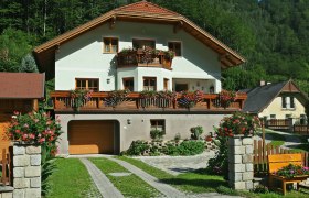 Privatzimmer Auer, © Roman Reichl Ein zweistöckiges Haus mit Holzbalkonen und Blumen, umgeben von grüner Landschaft.