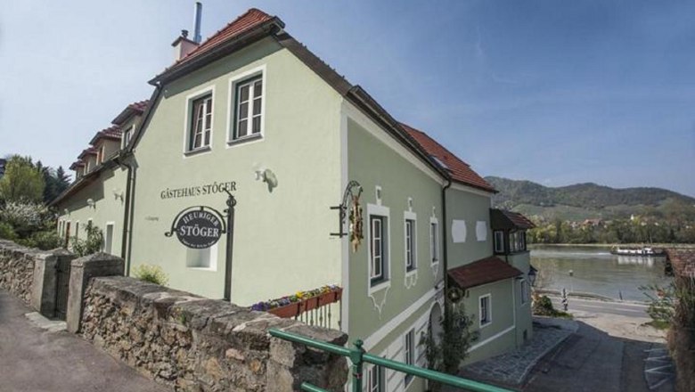 Winzerhof & Gästehaus Stöger, © Micheal Liebert Ein grünes Gästehaus mit roten Dächern am Flussufer, umgeben von Hügeln und blühenden Pflanzen.