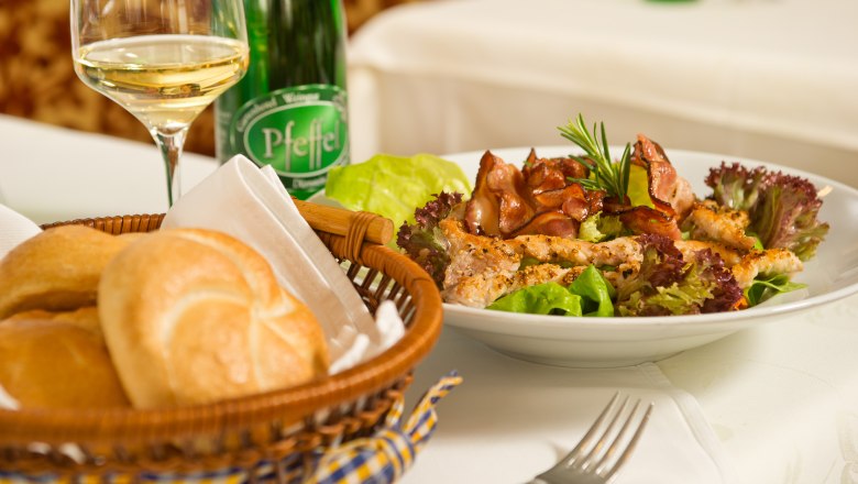 Vorspeise im Genuss.Restaurant, © Rita Newman Ein Teller mit Salat und Fleisch, ein Brotkorb und ein Glas Weißwein auf einem Tisch.