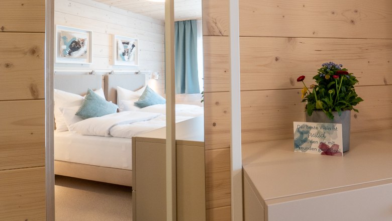 Gemütliches Ambiente, © freilich / IsaBella Fritsche Ein gemütliches Schlafzimmer mit Holzwänden, einem Bett mit blauen Kissen und einem kleinen Tisch mit einer Pflanze und einer Karte.