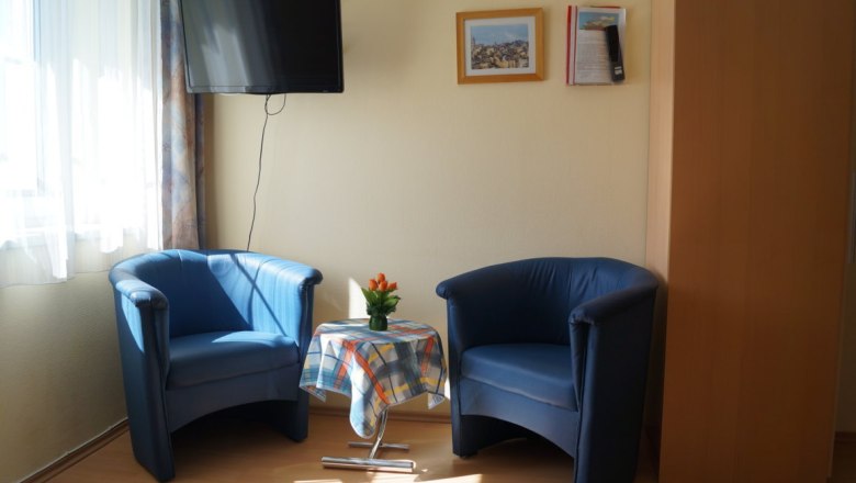 Sitzplatz im Buchenzimmer, © Reep Zwei blaue Sessel mit einem kleinen Tisch dazwischen, darauf eine Vase mit Blumen. Ein Fernseher hängt an der Wand, daneben zwei Bilder.