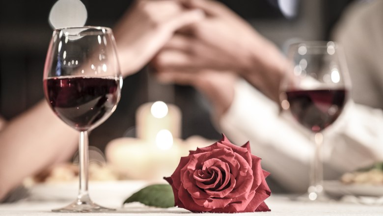 Romantikwochenende im Faulenzerhotel, © Karl Schweighofer Zwei Weingläser, eine rote Rose und ein Paar, das sich die Hände hält, bei Kerzenschein.