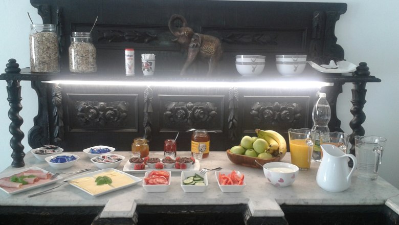 Frühstücksraum, © Ullram Frühstücksbuffet mit Obst, Aufschnitt, Käse, Marmelade und Getränken auf einem dekorativen Tisch.