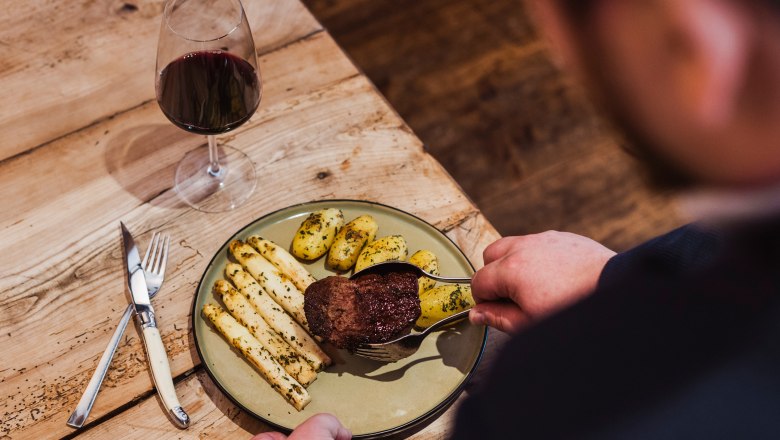 Rinderfilet am heißen Stein gebraten, © Niederösterreich Werbung/David Schreiber Ein Teller mit Rinderfilet, Spargel und Kartoffeln, daneben ein Glas Rotwein auf einem Holztisch.