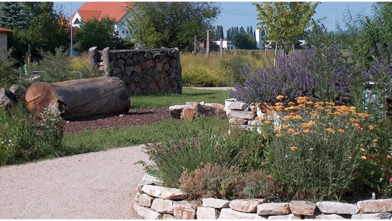 IGL Park, © Grünplan Ein gepflegter Garten mit Blumen, Bäumen und einem Holzstapel.