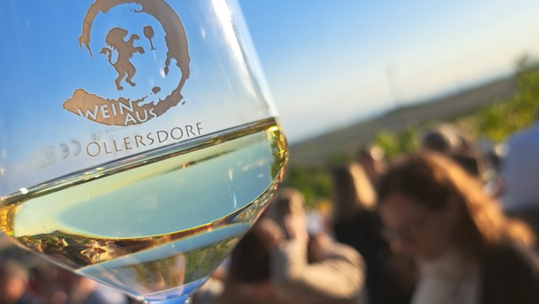 Tafeln im Weinviertel, © Weinviertel Tourismus GmbH Ein Weinglas mit der Aufschrift 'Wein aus Ollersdorf' wird in der Hand gehalten, im Hintergrund eine Gruppe von Menschen bei einem Outdoor-Event.