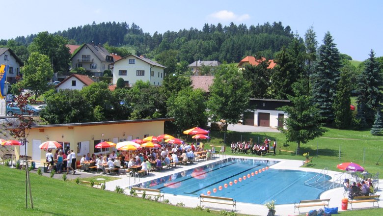 Freibad Els 01, © Ingrid Kleber Freibad mit Schwimmbecken, Sonnenschirmen und Menschen auf Liegestühlen, umgeben von grünen Hügeln und Häusern.