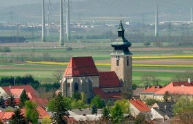 Kirche Pillichsdorf, © Thomas Falch Kirche in Pillichsdorf mit umliegenden Häusern und Windrädern im Hintergrund.