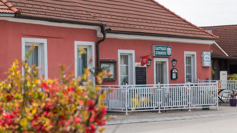 Gasthof Zivanovic, © Stadtgemeinde Traismauer Ein rosa Gebäude mit der Aufschrift 'Gasthaus Zivanovic', umgeben von einem weißen Zaun und Blumen im Vordergrund.