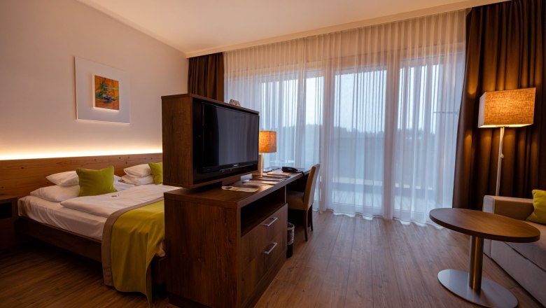 Hotel Schwarz Alm, © Schwarz Alm Modernes Hotelzimmer mit Doppelbett, Fernseher, Schreibtisch und großen Fenstern mit Vorhängen.