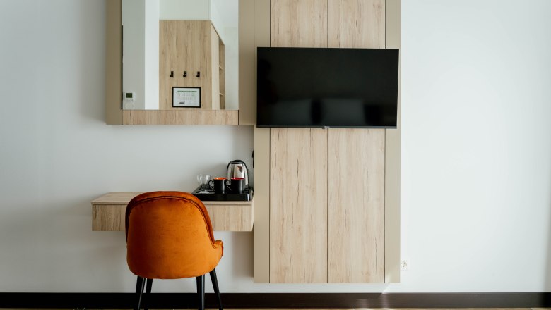 Hotel Lamede, © Hotel Lamede Modernes Hotelzimmer mit Holzmöbeln, einem Spiegel, einem Fernseher und einem orangefarbenen Stuhl.