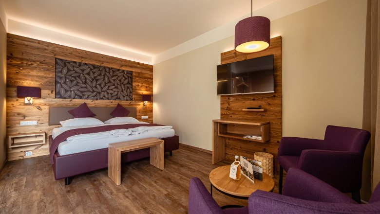 Bett in der Wies‘n, © Niederösterreich Werbung / Maximilian Pawlikowsky Gemütliches Hotelzimmer mit Holzwänden, Doppelbett, TV und lila Sesseln.
